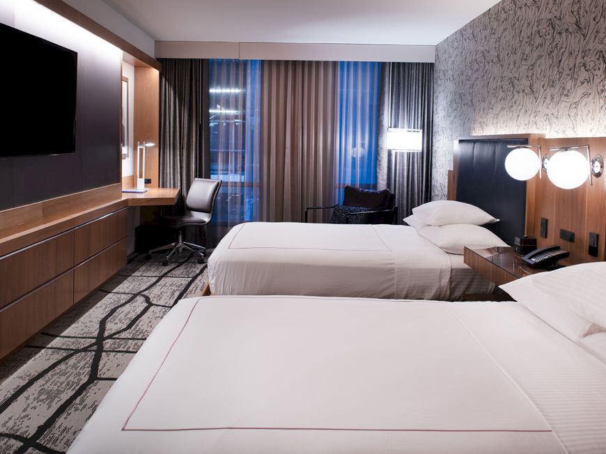 A hotel room features two neatly made beds, a flat-screen TV, a desk with a chair, modern decor, and large windows covered by curtains.