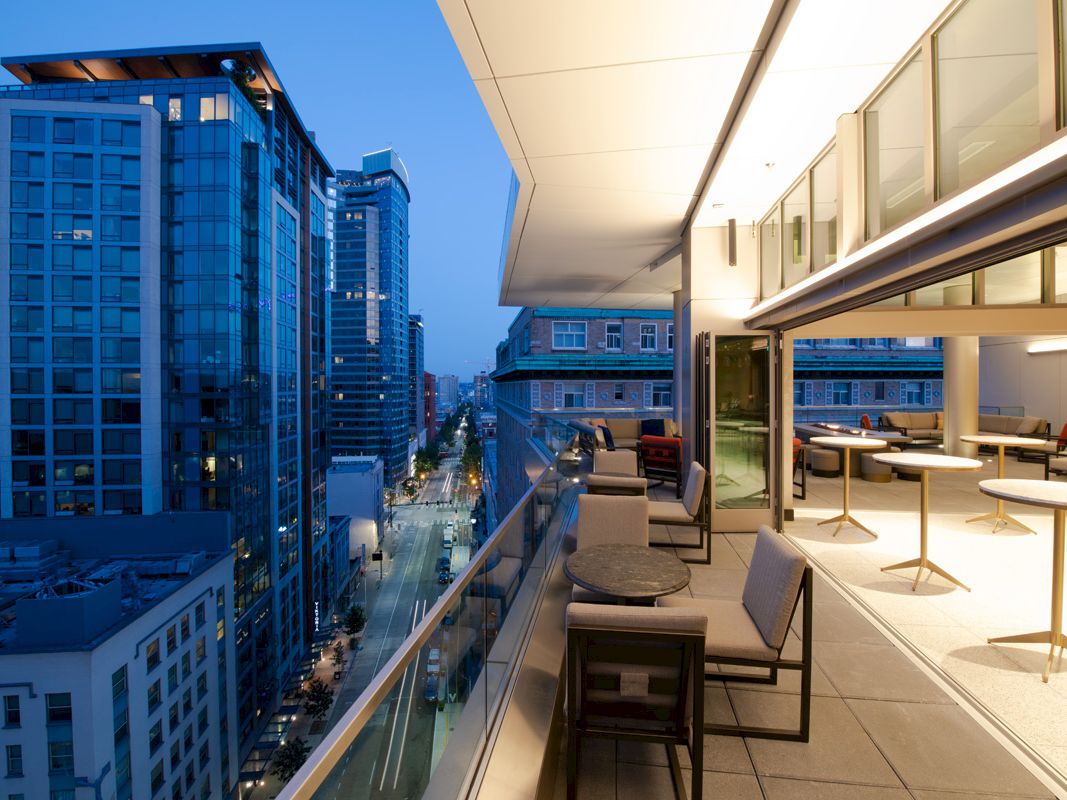 A modern cityscape at dusk with high-rise buildings. The foreground features a balcony with seating and tables, overlooking the city street below.