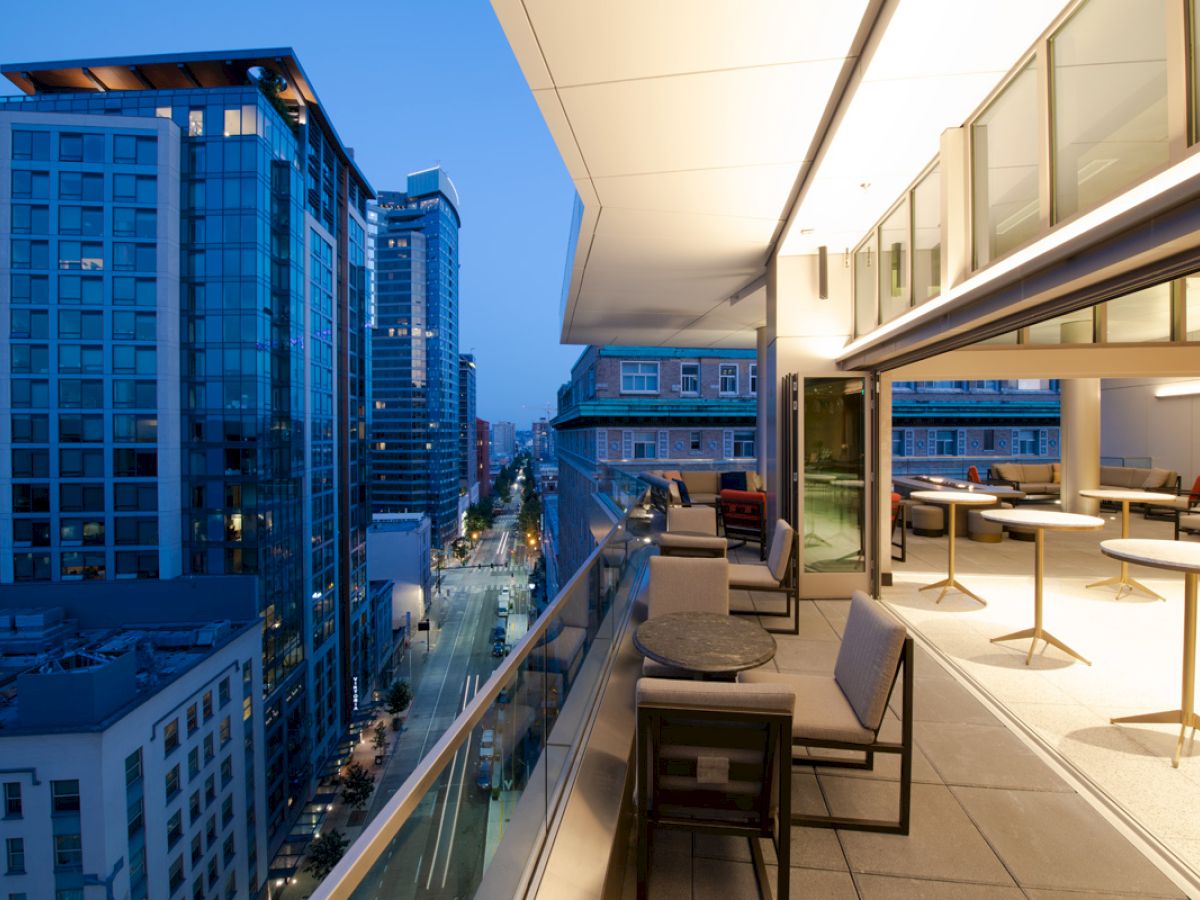 A rooftop balcony overlooking a cityscape at dusk, with outdoor seating and tables, and buildings lit up in the background.