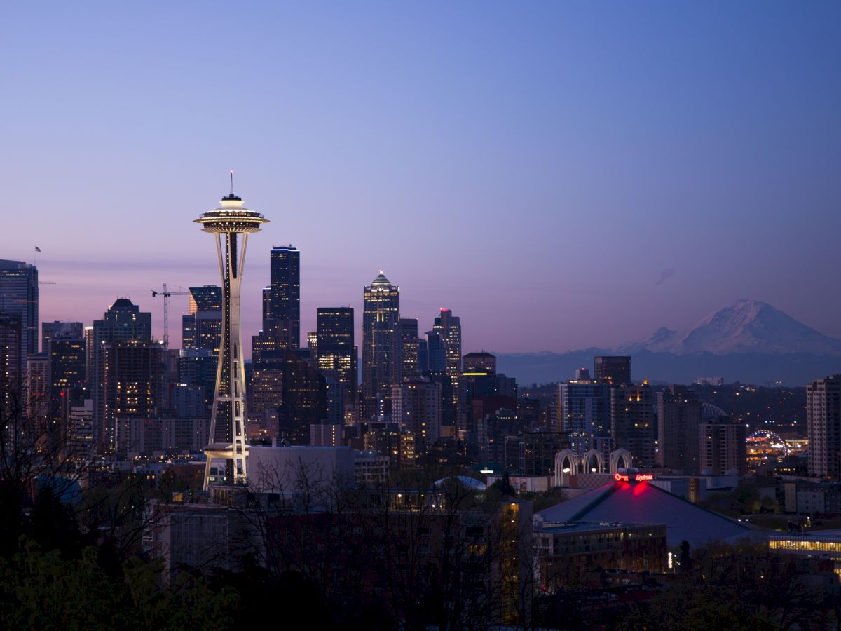 The image shows the Seattle skyline at twilight, featuring the iconic Space Needle with Mount Rainier in the background and city lights illuminating the scene.