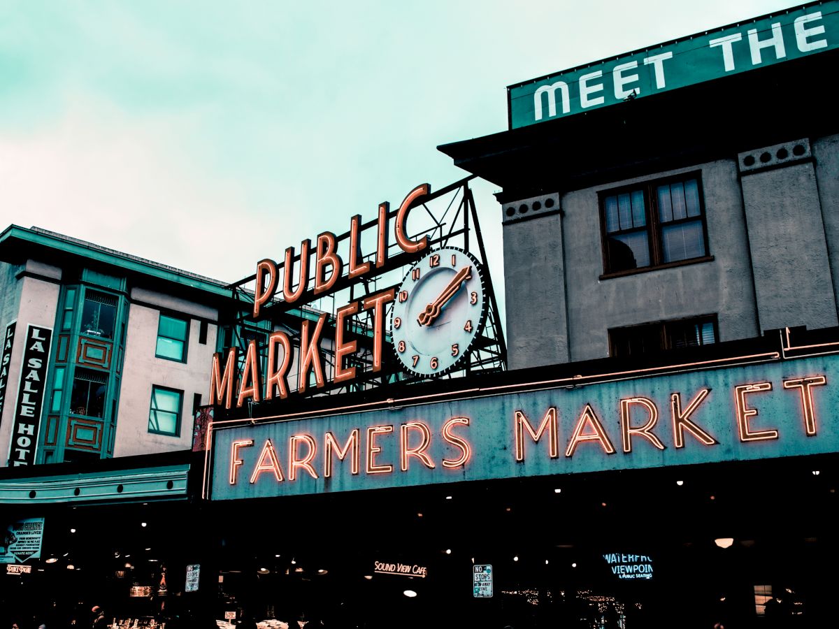 The image shows a neon sign for a public market and farmers market, with an iconic clock. Nearby buildings can be seen, including one labeled &ldquo;The&rdquo; hotel.