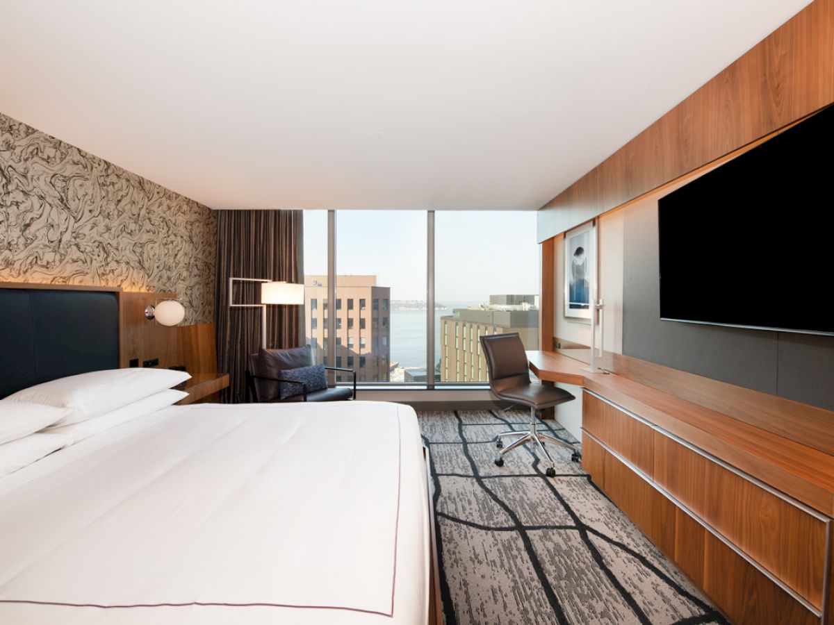 A modern hotel room with a large bed, a desk with a chair, a flat-screen TV, and a window view of buildings outside.