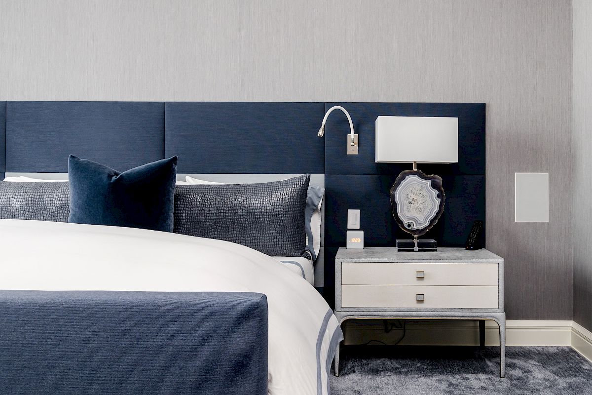 A modern bedroom featuring a blue headboard, white bedding, and a gray nightstand with a lamp and decorative object on a gray carpet.
