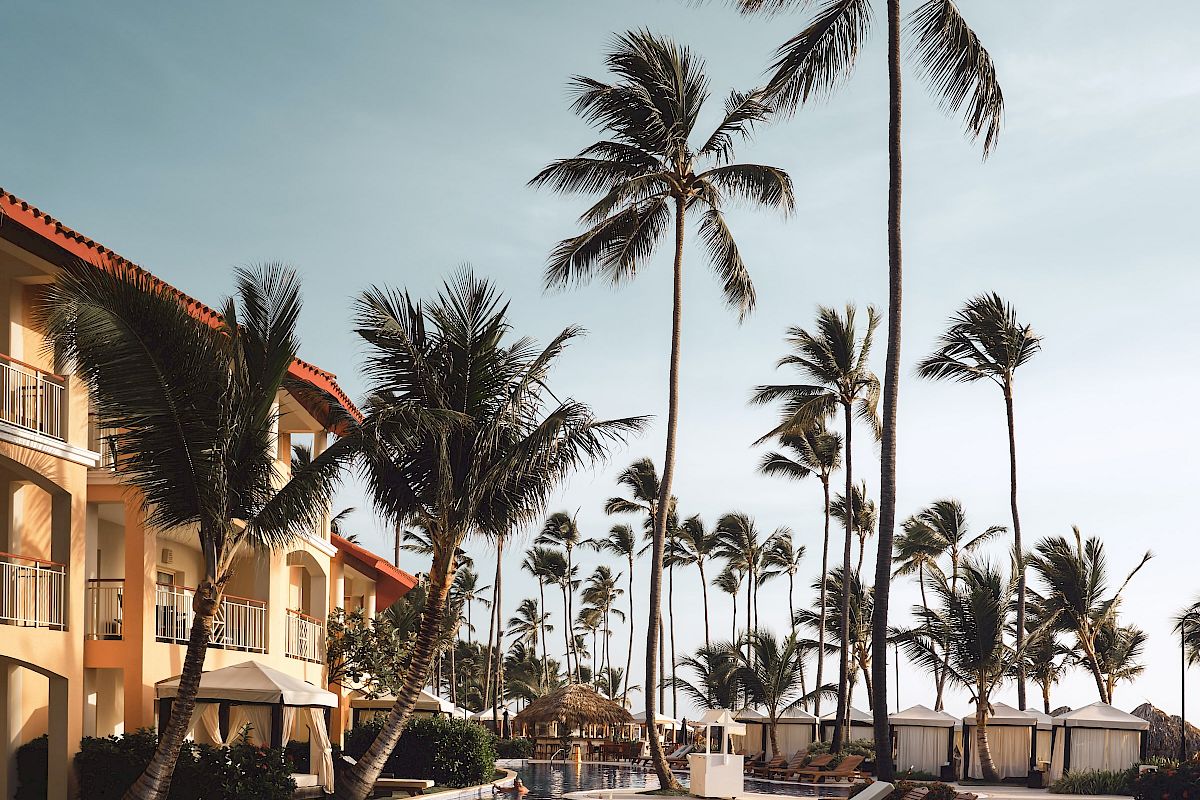 A luxurious poolside area with lounge chairs, surrounded by tall palm trees and a resort building in the background under a clear blue sky.
