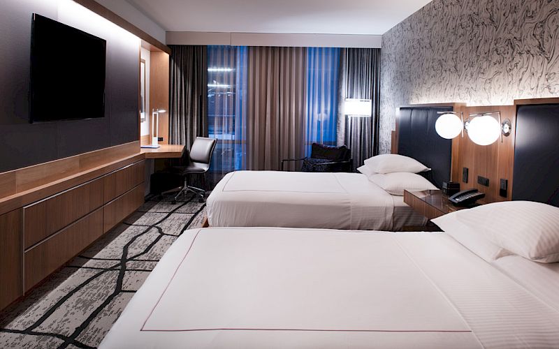 This image shows a modern hotel room with two neatly made beds, a flat-screen TV, a desk with a chair, and stylish decor on the wall.